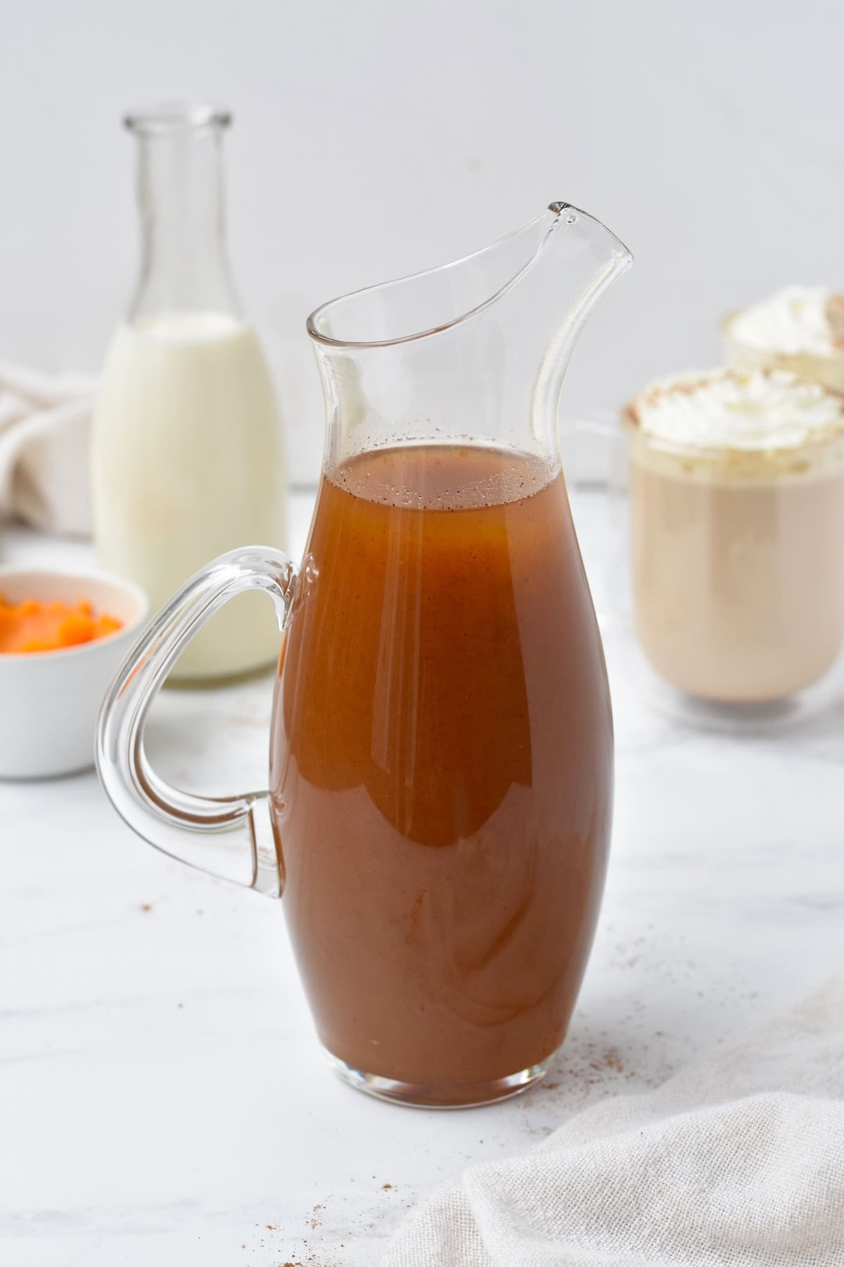 Pumpkin spice syrup in pouring jug in front of lattes.